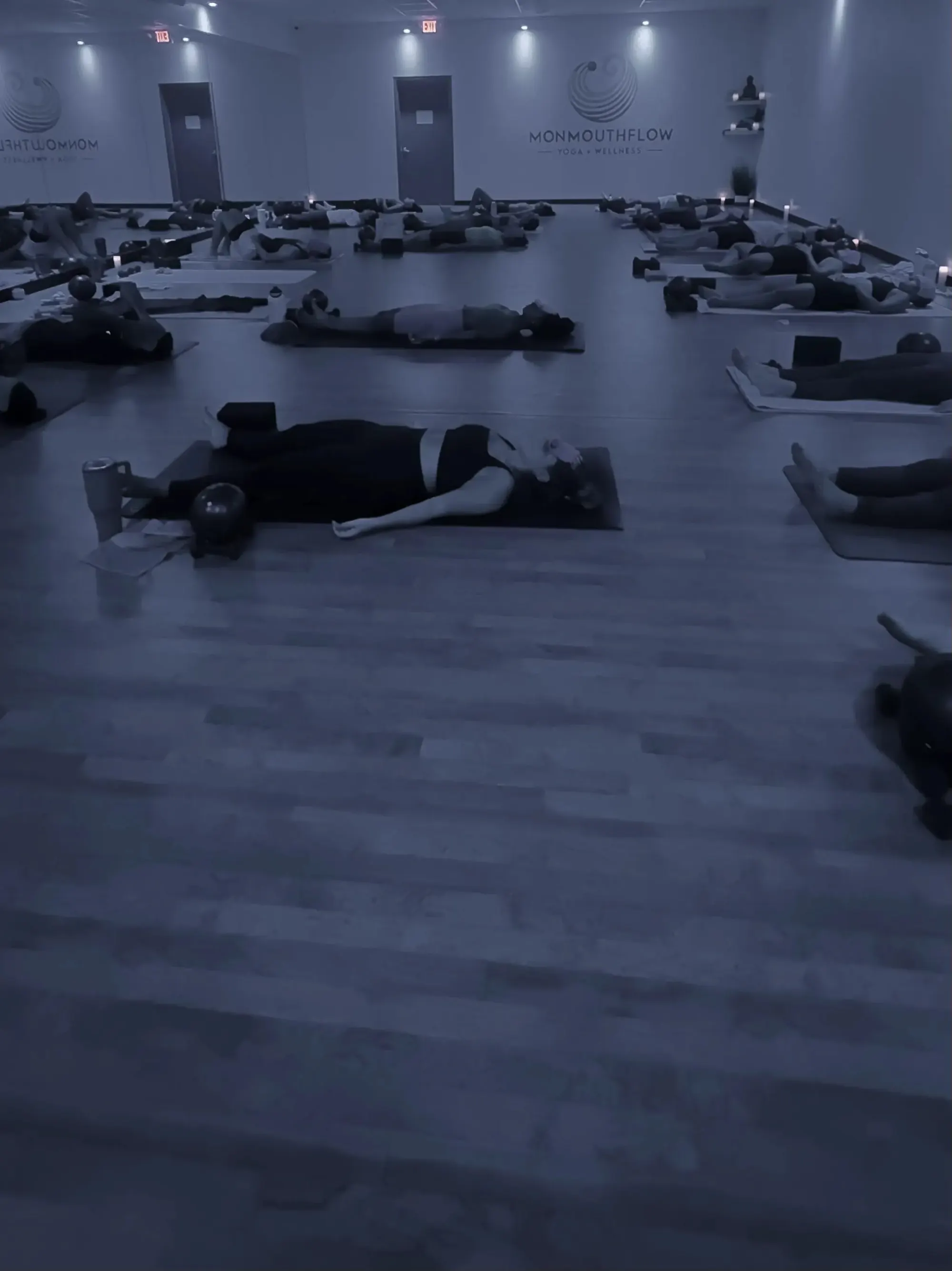 A candlelit class at MonmouthFlow resting in savasana on the studio floor, the MonmouthFlow logo on the far wall