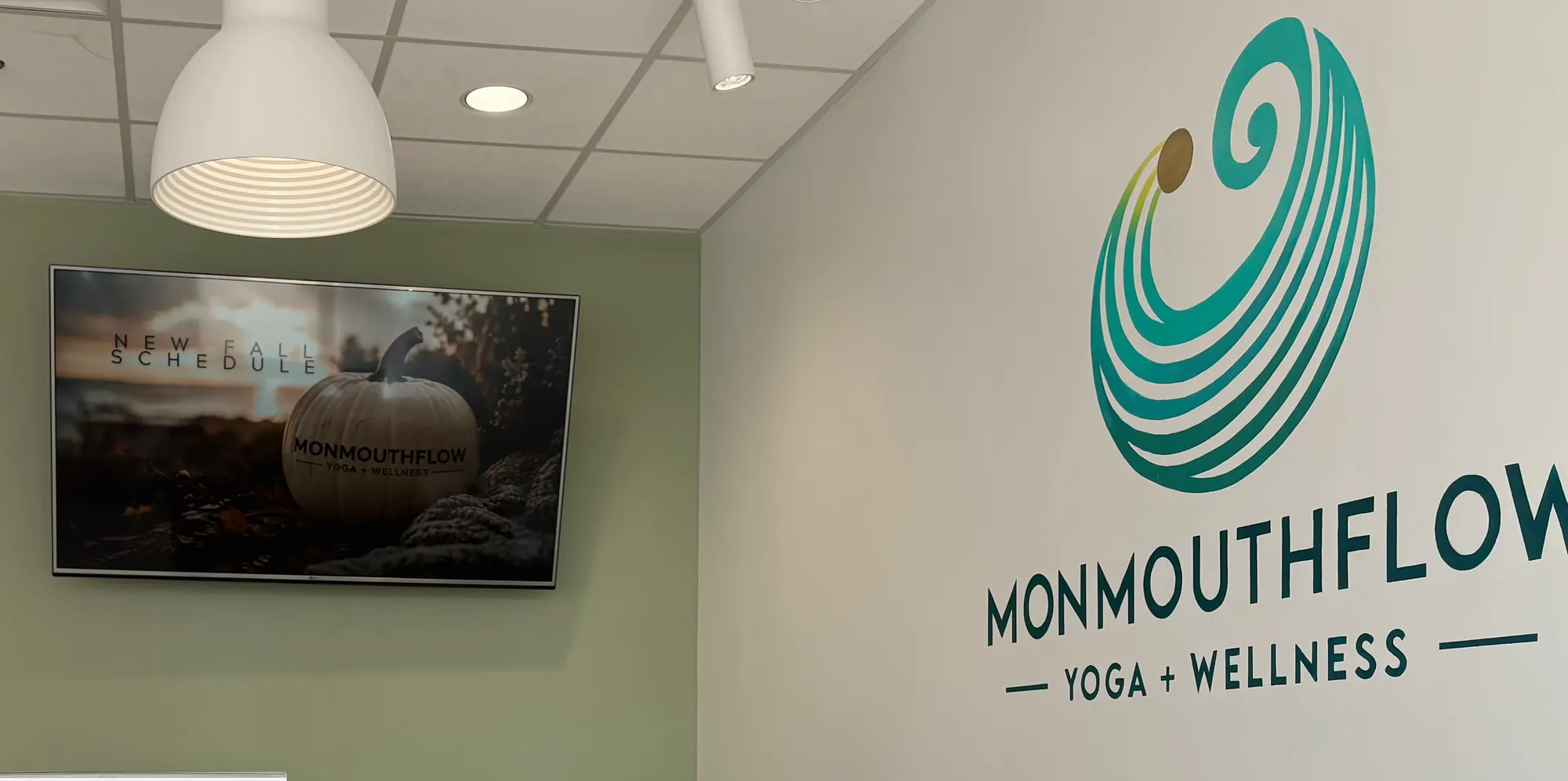 The MonmouthFlow Yoga + Wellness logo on the front desk wall, beside a screen showing the fall class schedule
